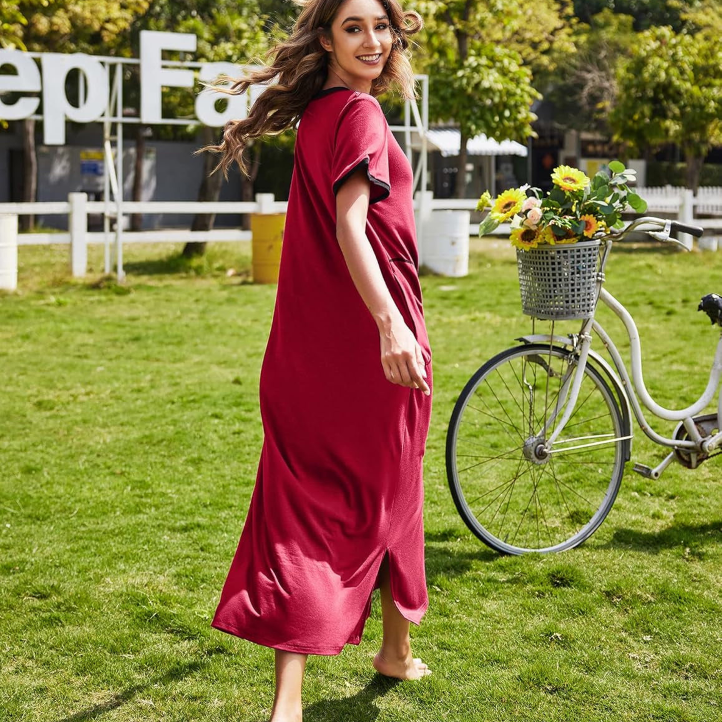 Woman in a red dress standing next to a bicycle with a basket of flowers on a grassy area.