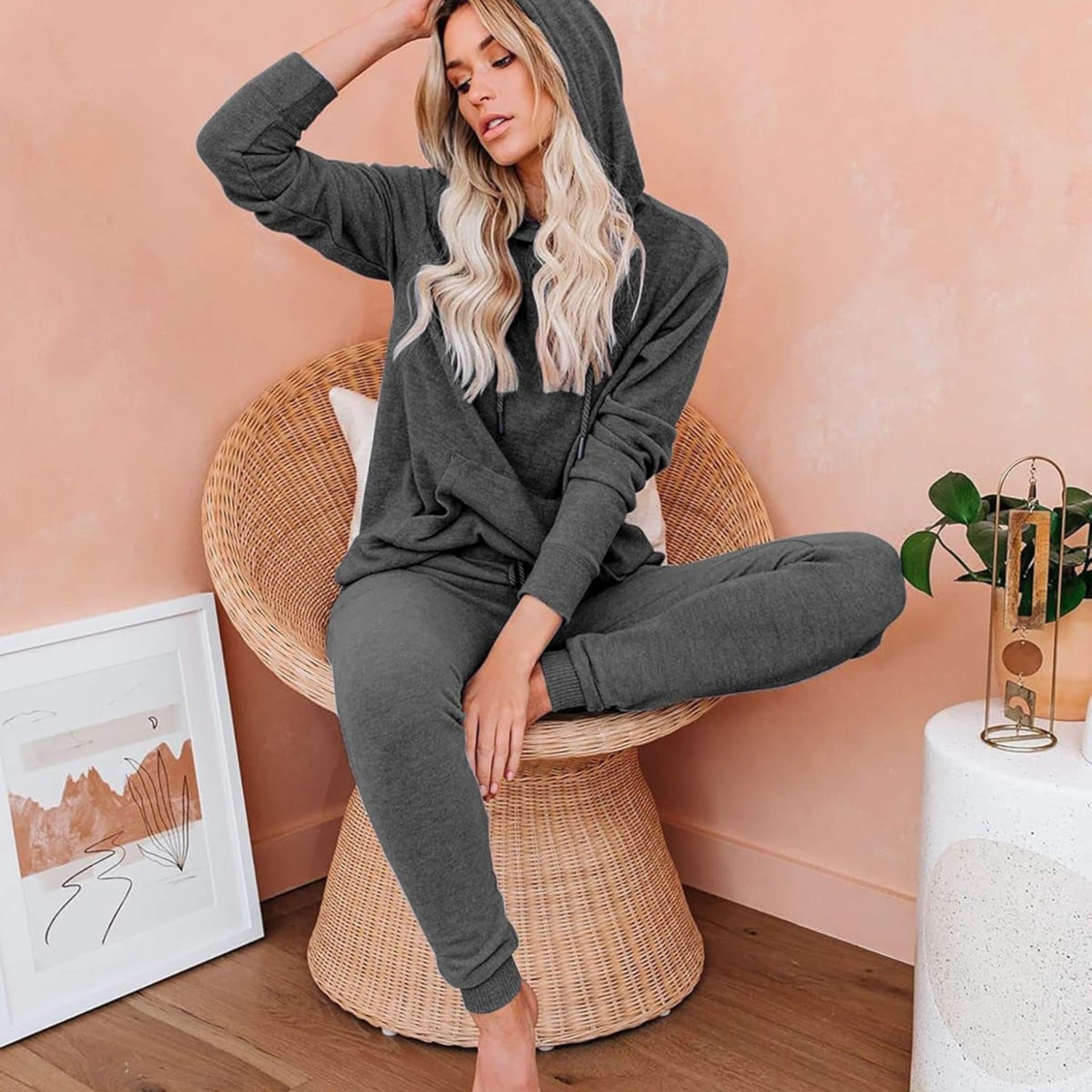Woman in a gray hoodie and pants sitting on a wicker chair against a pink wall.