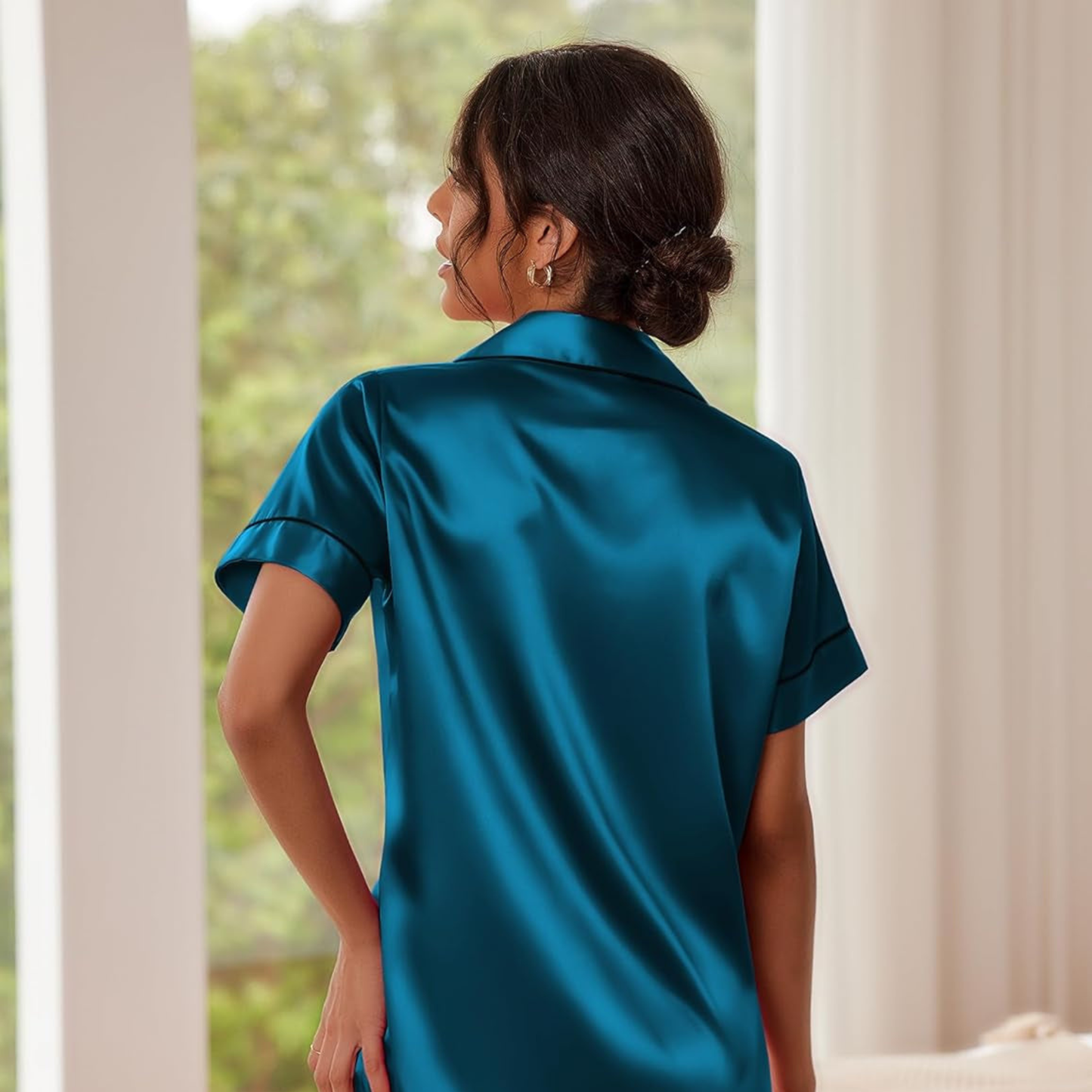 Woman wearing a teal satin shirt standing in a room with white curtains and greenery outside.